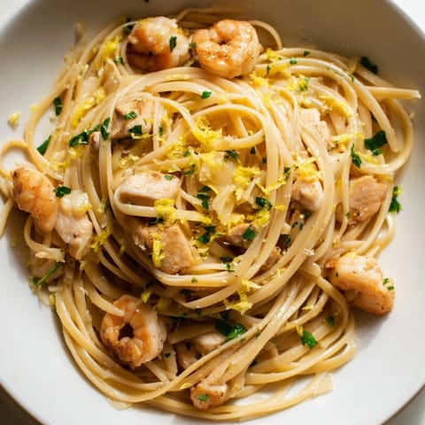 Plate of Lemon Garlic Shrimp & Chicken Pasta with linguine, golden chicken pieces, and pink shrimp in a glossy lemon-garlic butter sauce, garnished with fresh parsley and lemon wedges.  
