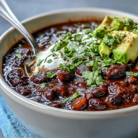 A steaming bowl of creamy Black Bean Soup garnished with sour cream, chopped cilantro, and diced avocado.