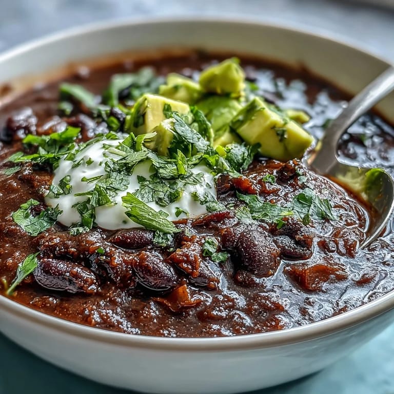 A ladle serving Black Bean Soup into a rustic bowl with lime wedges and a side of crusty bread.